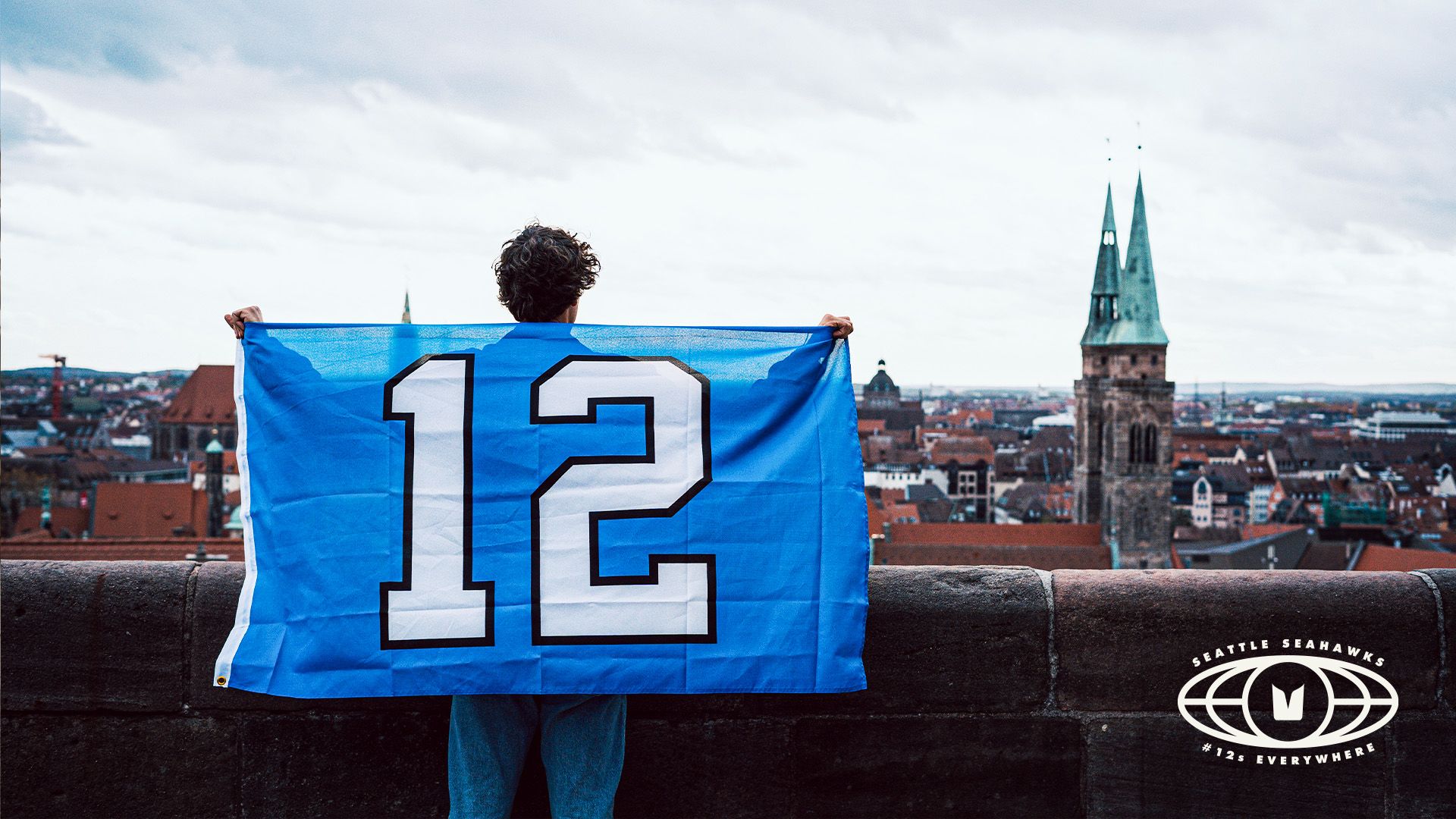A person stands facing a city, holding a blue 12 flag.